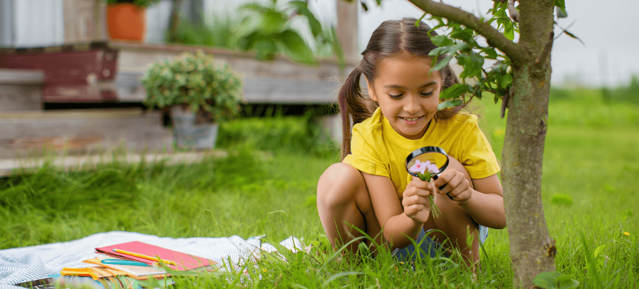 Child with magnifying glass examining pink flowers in grass.